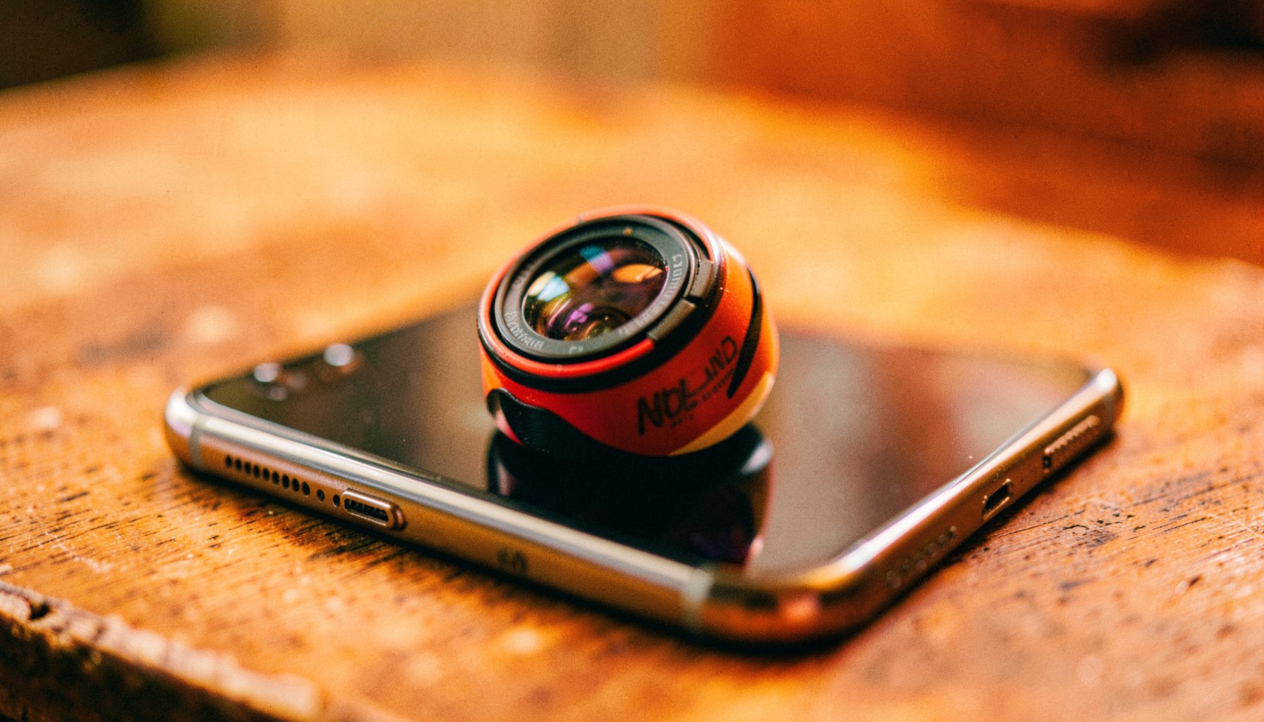 A close-up of an iPhone with a colorful Holga lens attachment resting on a wooden table, warm natural light, shallow depth of field